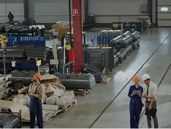 Workers in hard hats and coveralls standing on a factory floor with metal materials stacked on shelves