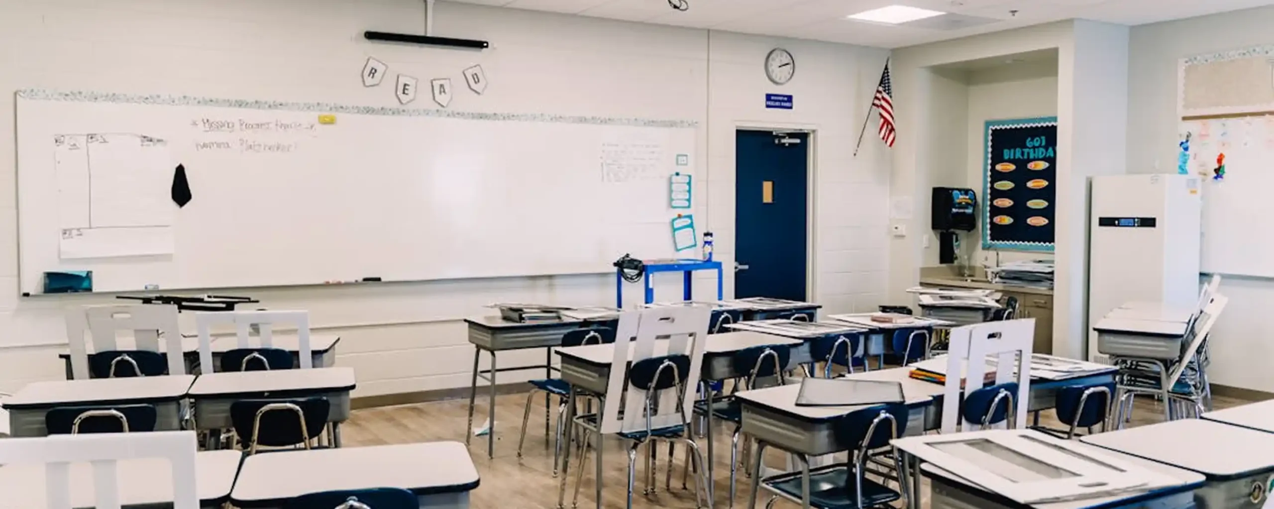 Empty classroom with rows of student desks, large whiteboard at front, wall clock and American flag